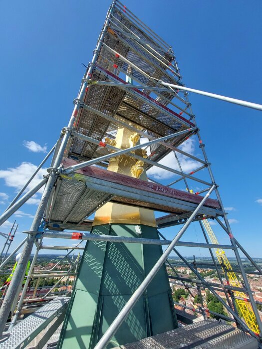 Byggnadsställning runt guldstaty på tak, med blå himmel och stadsvy i bakgrunden.