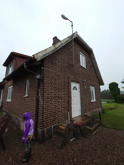 Tegelhus med stående gatlykta vid gavel, lösa kablar och stenhögar vid ingången. Regnig dag med barn i lila regnjacka.