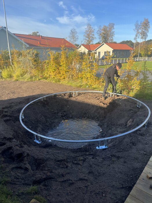 Man gräver runt en cirkulär ram i en lerig grop. I bakgrunden syns hus med röda tak.