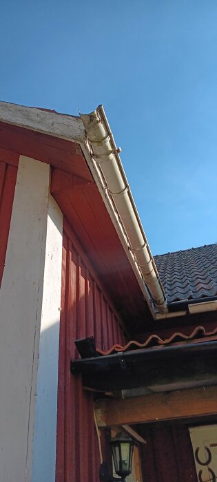 Bild av ett rött hus med vita knutar och ett gammalt vitt stuprör. Taket har bruna takplattor och ljusblå himmel syns i bakgrunden.