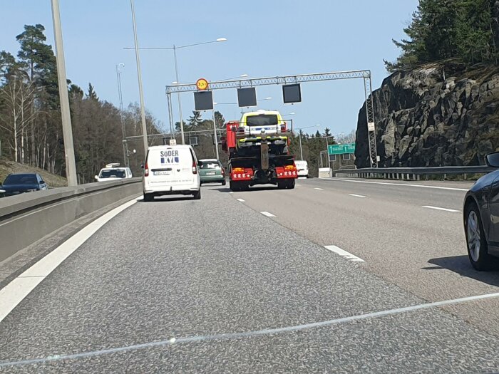 Biltransport med lastbil på motorväg, omgiven av andra bilar och vägskyltar, passerar skogklädda klippor under en klar himmel.