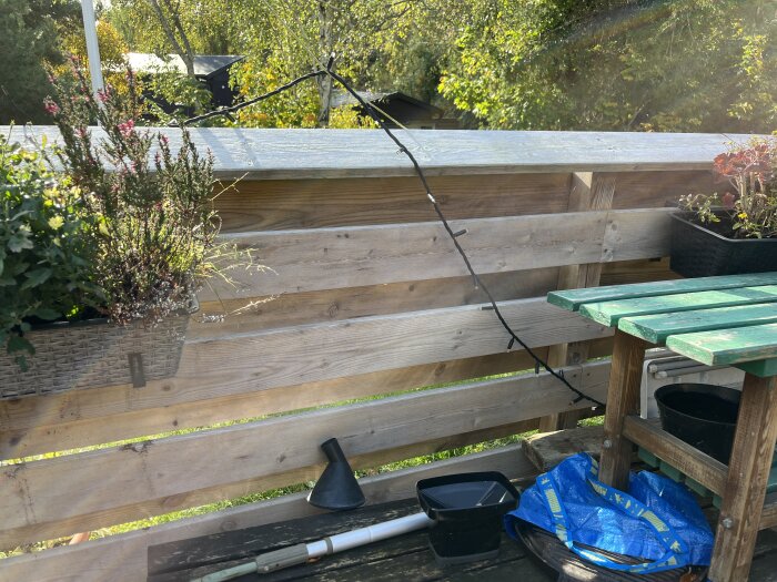 Balkong med trästaket, dekorerad med blommor i krukor och ljusslinga. Grönt bord och trädgårdsverktyg syns, med skog i bakgrunden.