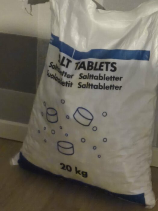 En 20 kg påse med salttabletter, etikett på flera språk med bilder av tabletter, står på ett golv.