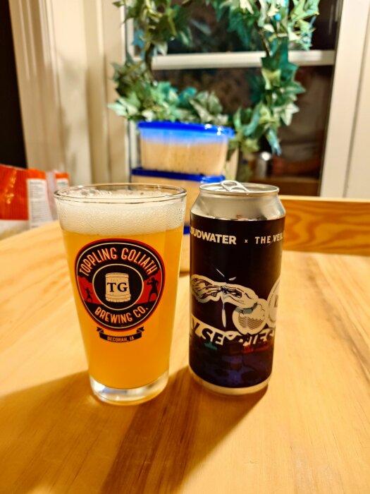 Ett ölglas med logotyp från Toppling Goliath Brewing Co och en ölburk från Cloudwater på ett bord, med en grön växt i bakgrunden.