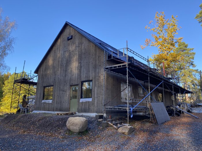 Trähus med grånad fasad behandlad med järnvitriol, omgiven av byggställningar och höstlandskap.