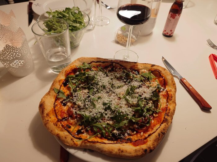 Hemgjord pizza toppad med ruccola, parmesan och ringlad balsamico. Pizzan serveras med ett glas rött vin och sallad i bakgrunden.