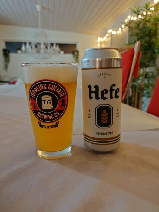 Ett glas fyllt med veteöl och en burk Hefeweizen öl står på ett bord i ett rum med dekorativa ljus i bakgrunden.