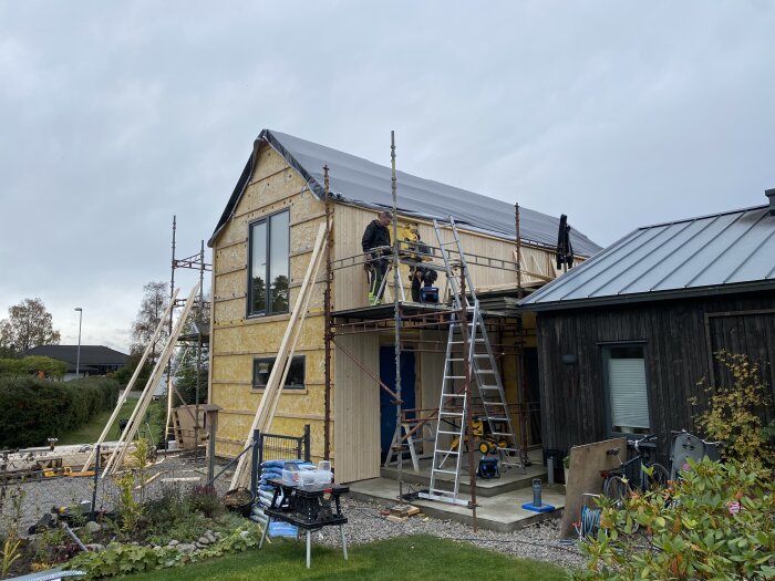 Renoveringsprojekt med byggnadsställning och granpanel monterad på en tillbyggnad, arbetare och verktyg på plats.