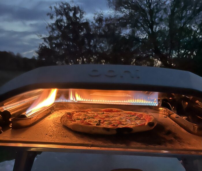 Pizza i en ugn med brända kanter och lågor i bakgrunden, kvällstid utomhus.