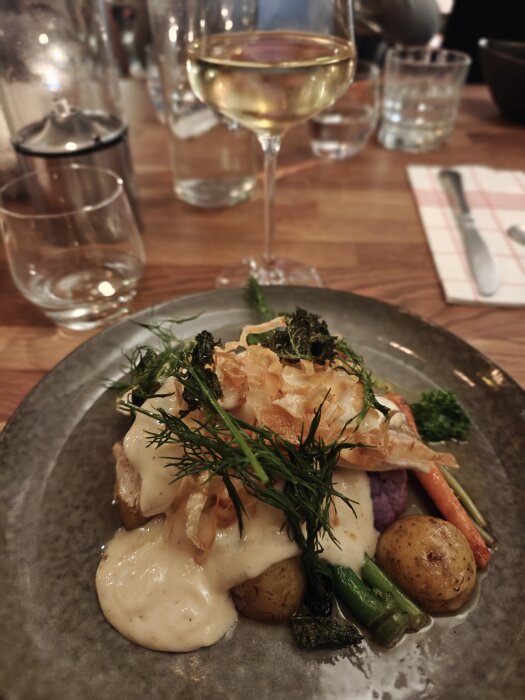 En tallrik med potatis, grönsaker, krämig sås och dill på en restaurang i Örebro, med ett glas vin bredvid.