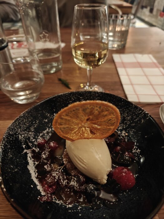 En dessert med glass, hallon och en torkad apelsinskiva på en tallrik, samt ett glas vitt vin på ett träbord.