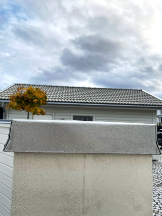 Plåtbeklädnad på ett plank med synliga bucklor, framför en vit byggnad och molnig himmel.