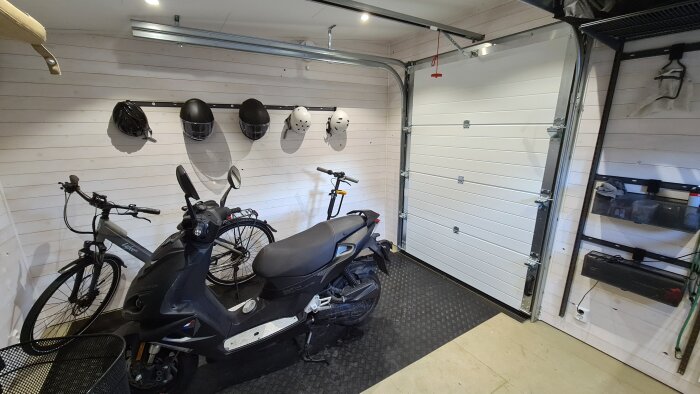 Inuti en isolerad garagebod med cykel och moped, hjälmar på en vägg, och en stängd vit garageport.