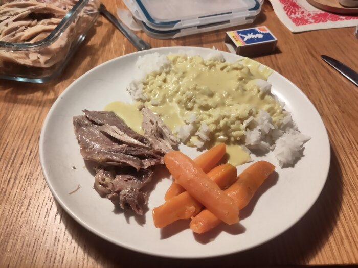 Tallrik med kokt tupp i currysås, ris och morötter på ett bord. I bakgrunden en glasskål med mer kött.