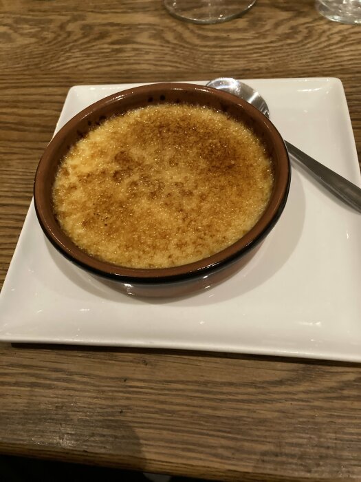 Créme brulée i brun ugnsform på kvadratisk vit tallrik med sked, serverad på ett träbord.