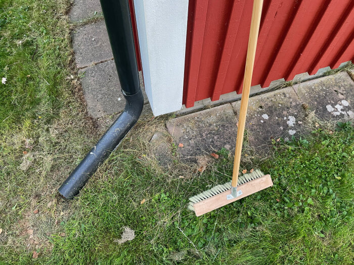 Svart stuprör avslutas nära husgrund bredvid en röd vägg och en borste står lutad mot väggen. Gräs och plattor syns på marken.