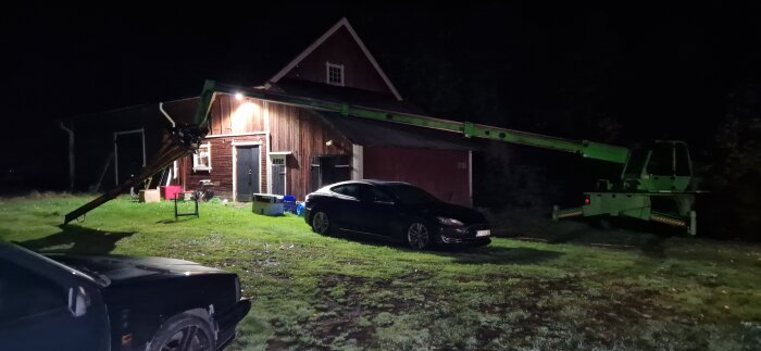 Nocturnal scene with a garage, a black car parked in front, and a green construction crane extending towards the roof. Lit by outdoor lights.