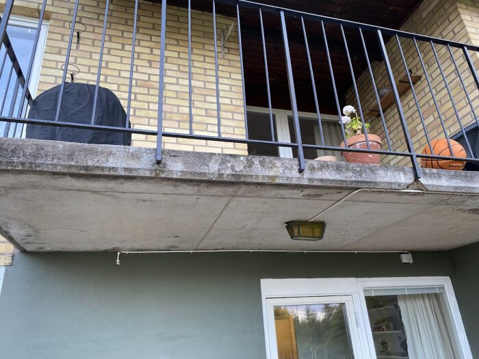 Balkong med svart grillöverdrag, blomkruka och basketboll, försedd med metallräcke. Betongen visar tecken på slitage. Brunt tegelfasadhus.