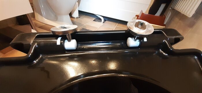 Närbild av en svart toalettsits med monteringsfästen i vitt och metall, utan synliga justeringsmekanismer.