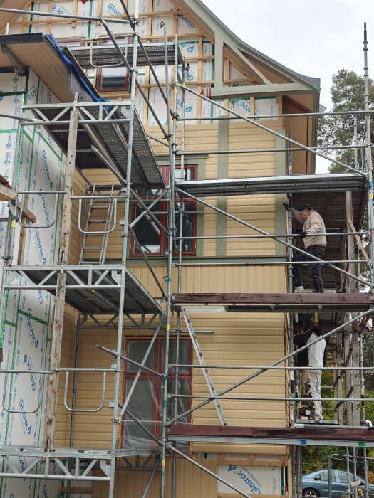 Byggställningar framför ett hus med pågående fasadrenovering, karvsnitt på sidan av fönster och arbetare som målar och installerar paneler.