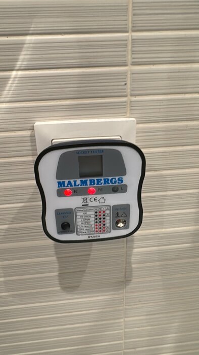 Elektrisk uttagstestare från Malmbergs ansluten till ett vägguttag med två röda lysdioder tända, som visar fas/nolla-status.