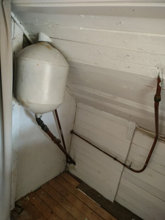 Gammalt expansionskärl och rör i hörn med träpanel på vägg och golv.