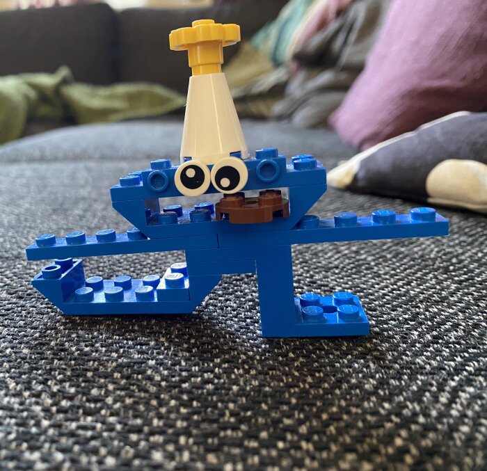 Legofigur med blå klossar och runda ögon, formad som ett djur eller monster, stående på ett soffbord med kuddar i bakgrunden.