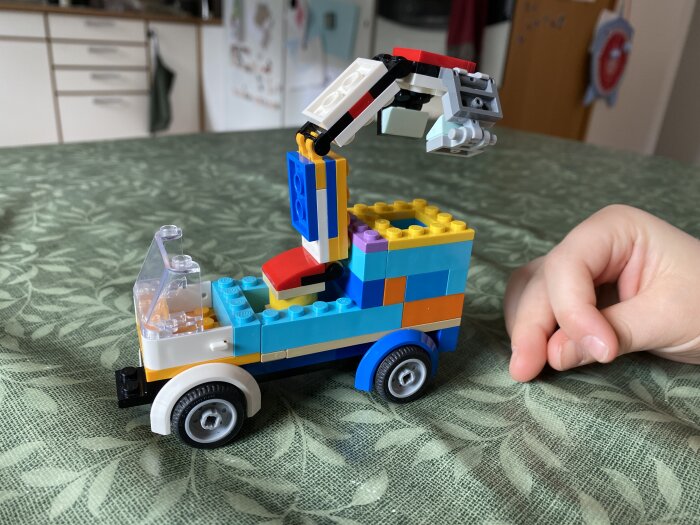 Liten Lego-bil med kran, byggd av färgglada klossar, placerad på ett grönt bord. Ett barns hand syns bredvid bilen.