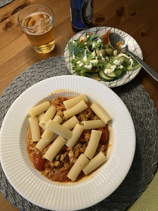 Tallrik med pollognese, rigatoni-pasta och tomatsås, bredvid står öl, sallad, och ett glas dryck på ett träbord.