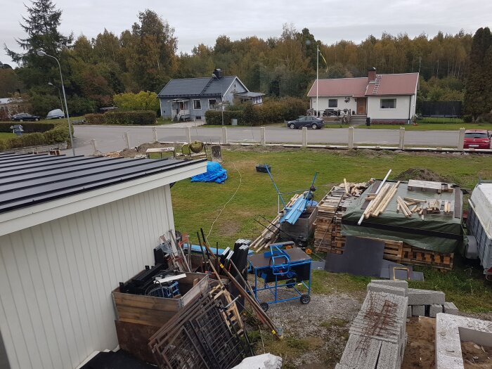 Byggmaterial och verktyg staplade utomhus på en gräsmatta, med synliga gjutna stolpar och ett bostadsområde i bakgrunden.