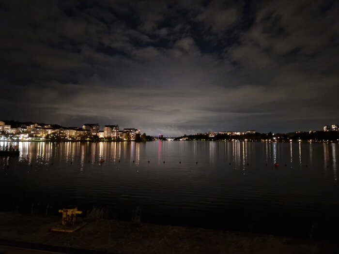 Stadslandskap vid vatten i nattljus med reflektioner från upplysta byggnader och molnig himmel.