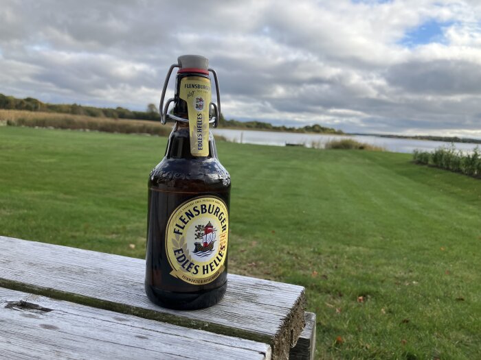 Flaska Flensburger öl på träbord utomhus med gräsfält och sjö i bakgrunden under molnig himmel.