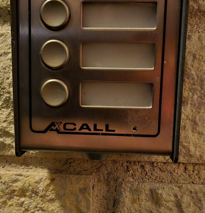 Metallisk porttelefon av äldre modell med tre knappar och namnskyltar på en tegelvägg, märkt "XCALL". Diskuteras för kodlåsinstallation.