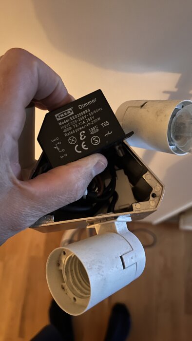 Hand som håller en IKEA-dimmer med specifikationer för utbyte av gamla glödlampsdimmers, bredvid en vit lamphållare med vridskruv.