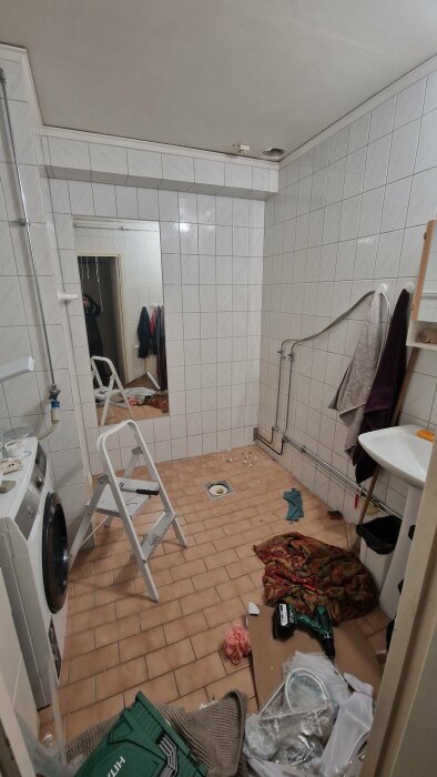 Badrum under renovering med vitkaklade väggar, brun klinkergolv, stege, tvättmaskin och verktyg utspridda på golvet. Hål för fläkt synligt i taket.