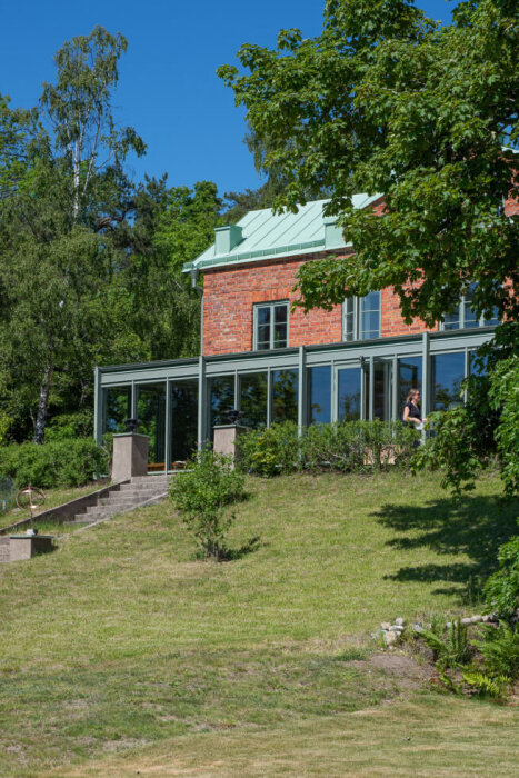 Tegelhushus med en tillbyggnad i glas och metall på bottenvåningen, omgiven av grönska och en gräsmatta.