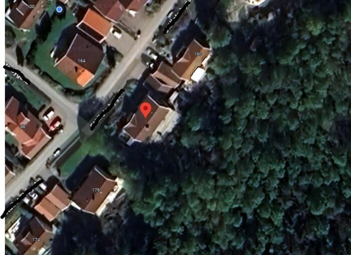 Satellitbild över ett bostadsområde med flera hus, vägar och skog, röd markör vid ett hus.