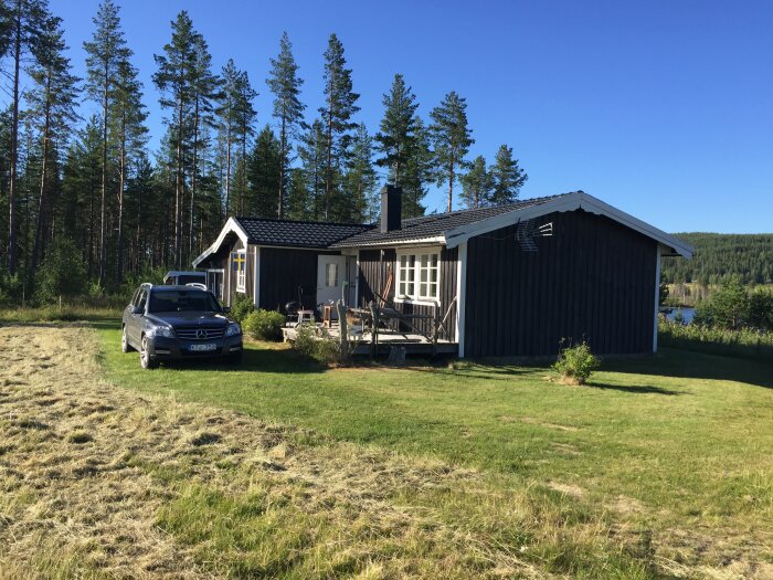 Fritidshus med garage på landsbygden, omgiven av träd, under klarblå himmel. Parkerat fordon vid sidan och sjö i bakgrunden.