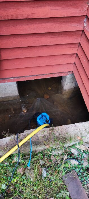Grundvattenproblem under hus med röd träpanel, visar en blå vattenpump i en översvämmad krypgrund.