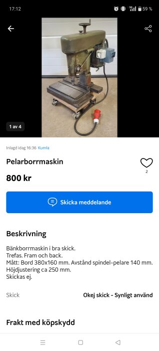 Gammal pelarborrmaskin i metall på hjulförsedd basplatta, med strömkabel, visas inomhus framför ljus vägg.