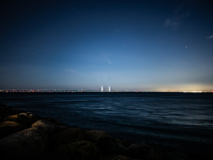 Nattbild av Öresundsbron över vattnet med stjärnklar himmel och ljus längs horisonten.