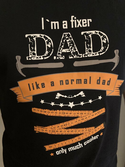 T-shirt med text "I'm a fixer dad like a normal dad only much cooler", dekorerad med hammare och måttband i orange och vit.