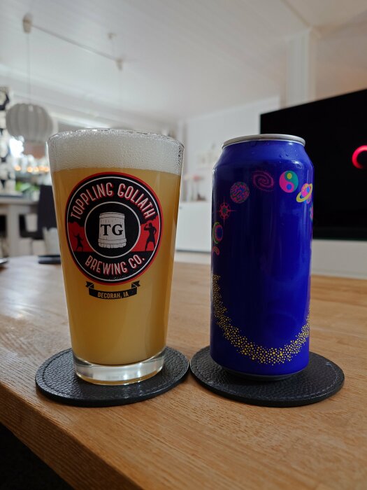 Ett glas öl med Toppling Goliath-logotyp bredvid en blå ölburk på ett bord i ett vardagsrum.