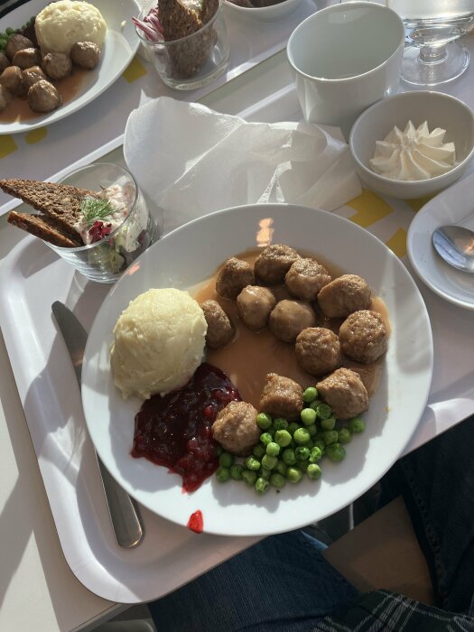 Tallrik med IKEA-köttbullar, potatismos, ärtor och lingonsylt. I bakgrunden en glasbägare med kavring och skagenröra.