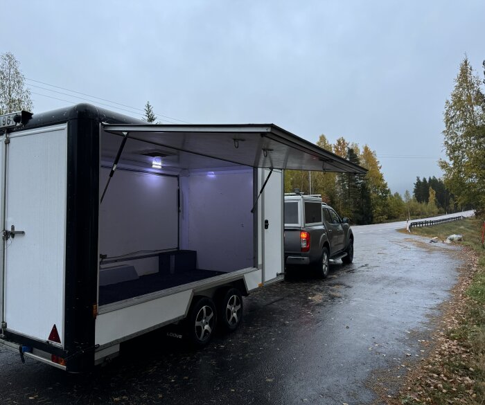 Släpvagn kopplad till en bil på en landsväg; släpet är öppet och inrett med belysning, planeras för omklädning och förvaring av verktyg och snöskotrar.