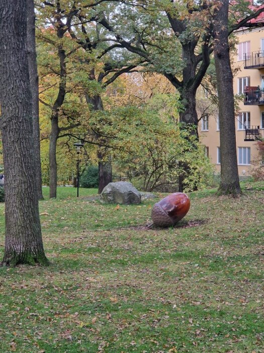 Park med stora träd och ett ekollonformad skulptur på gräsmattan, med flervåningshus i bakgrunden.