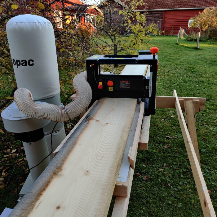 Vevor-planhyvel som hyvlar en bred planka utomhus, kopplad till en spånutsugningsapparat, omgiven av grönskande trädgård.