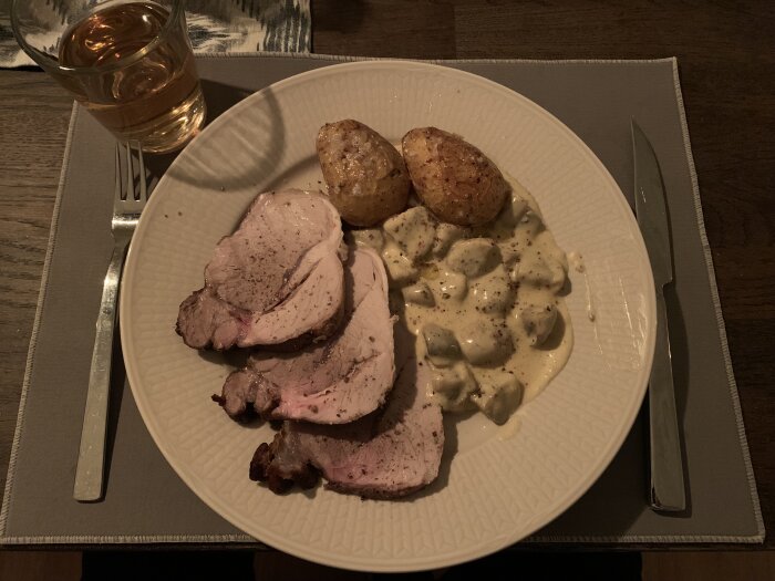 Tallrik med skivat kött, rostad potatis och gräddig svampsås, samt glas med dryck på träbord.