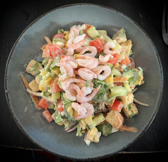 Räksallad med grönsaker och krutonger på en tallrik, med ingredienser som tomat, gurka och majs, toppat med skalade räkor.