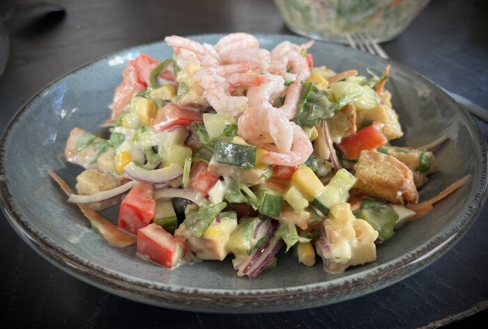 Räksallad med grönsaker, krutonger och dressing på en grå tallrik.